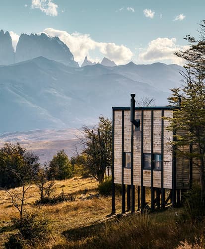 Modern cabin on stilts set in a grassy landscape with trees, overlooking dramatic mountain peaks under a clear sky.