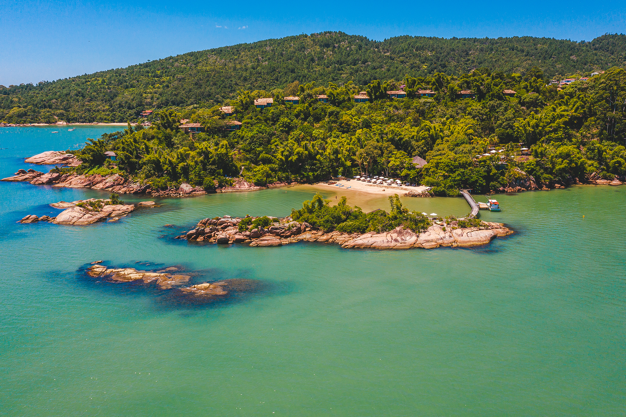 Aerial view of a lush tropical island with turquoise waters, rocky shores, beach loungers, and hillside villas.
