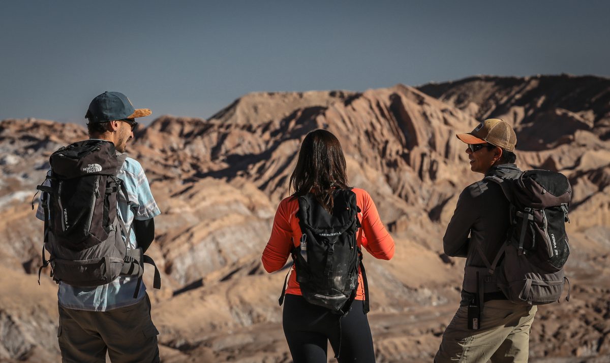 Three hikers with backpacks standing on a rocky overlook, facing layered desert mountains under a clear sky.