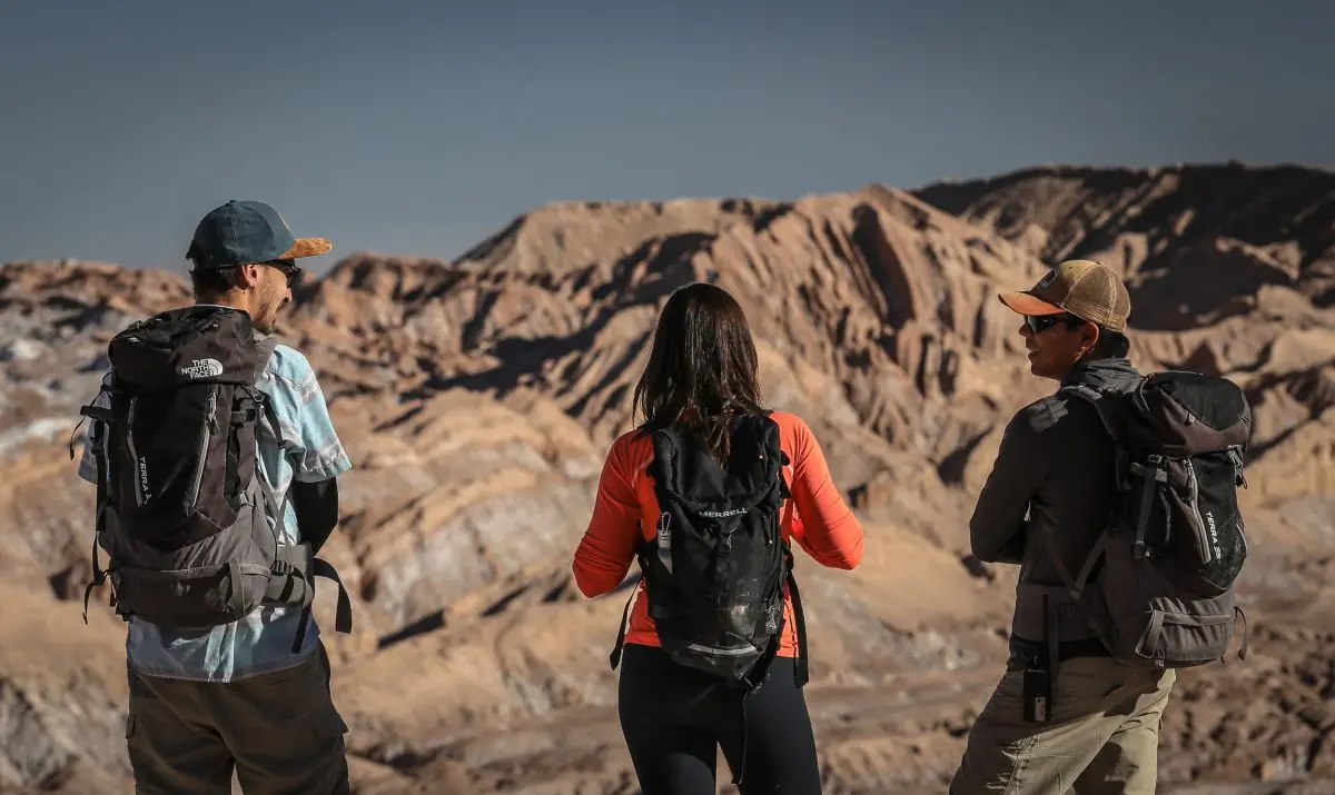 Three hikers with backpacks standing on a rocky overlook, facing layered desert mountains under a clear sky.