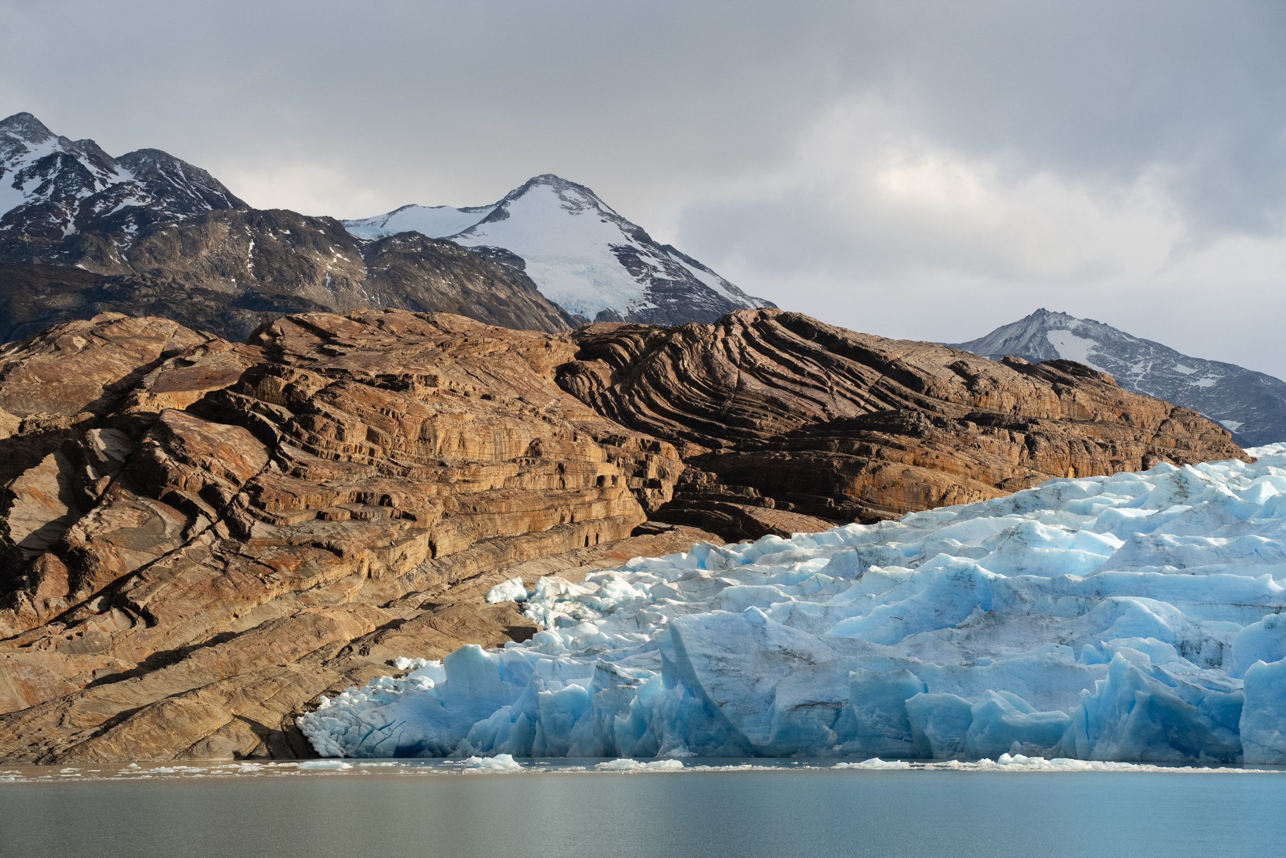 Blue glacier meets rocky mountains and calm water, with snow-capped peaks under a soft, overcast sky.
