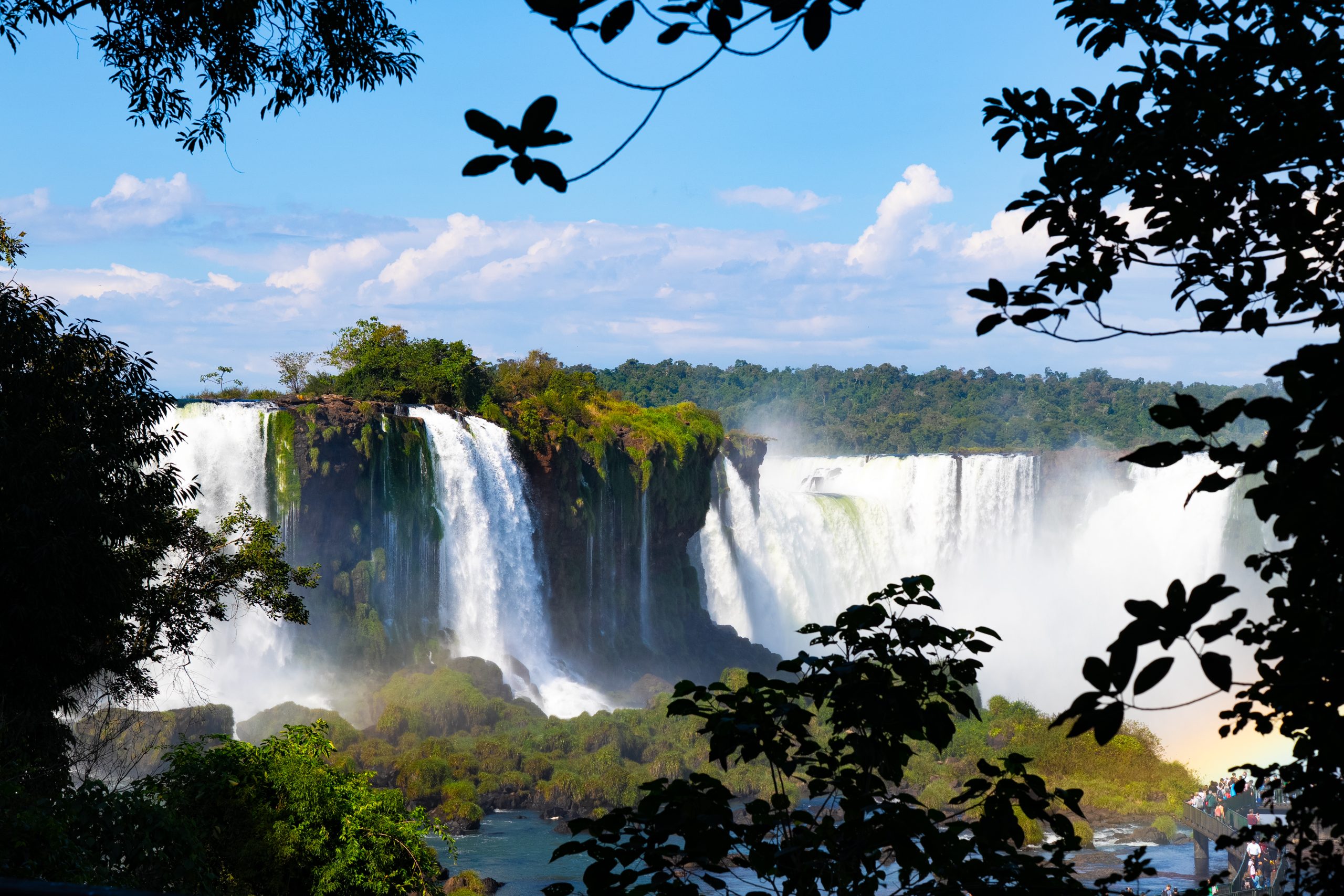 Wide waterfall cascading over lush cliffs, framed by leafy trees under a bright blue sky.