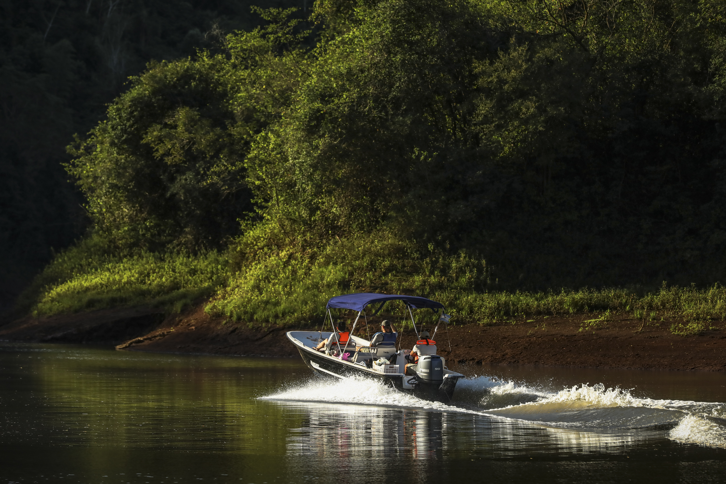 Small motorboat with passengers wearing life vests speeds along a calm river bordered by dense green trees.