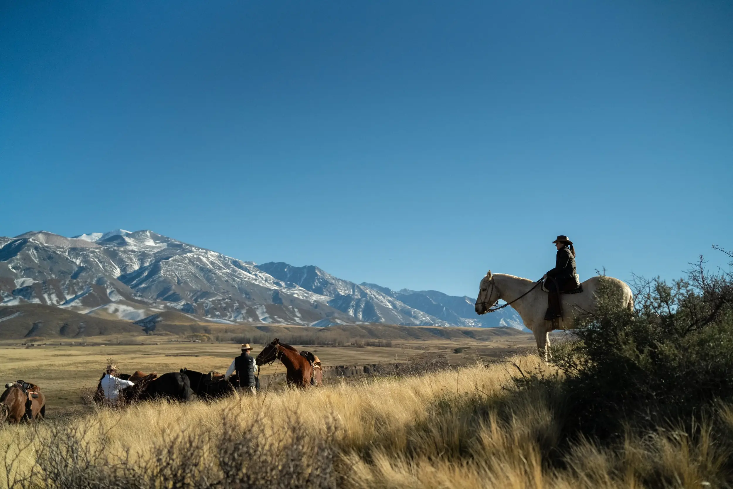 Riders On Horseback Crossing Open Grassland With Snow-Capped Mountains In The Distance Under A Clear Blue Sky.