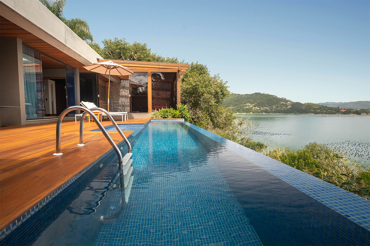 Infinity pool beside a modern villa overlooking a calm bay, with wooden deck, lounge chairs, and clear blue sky.