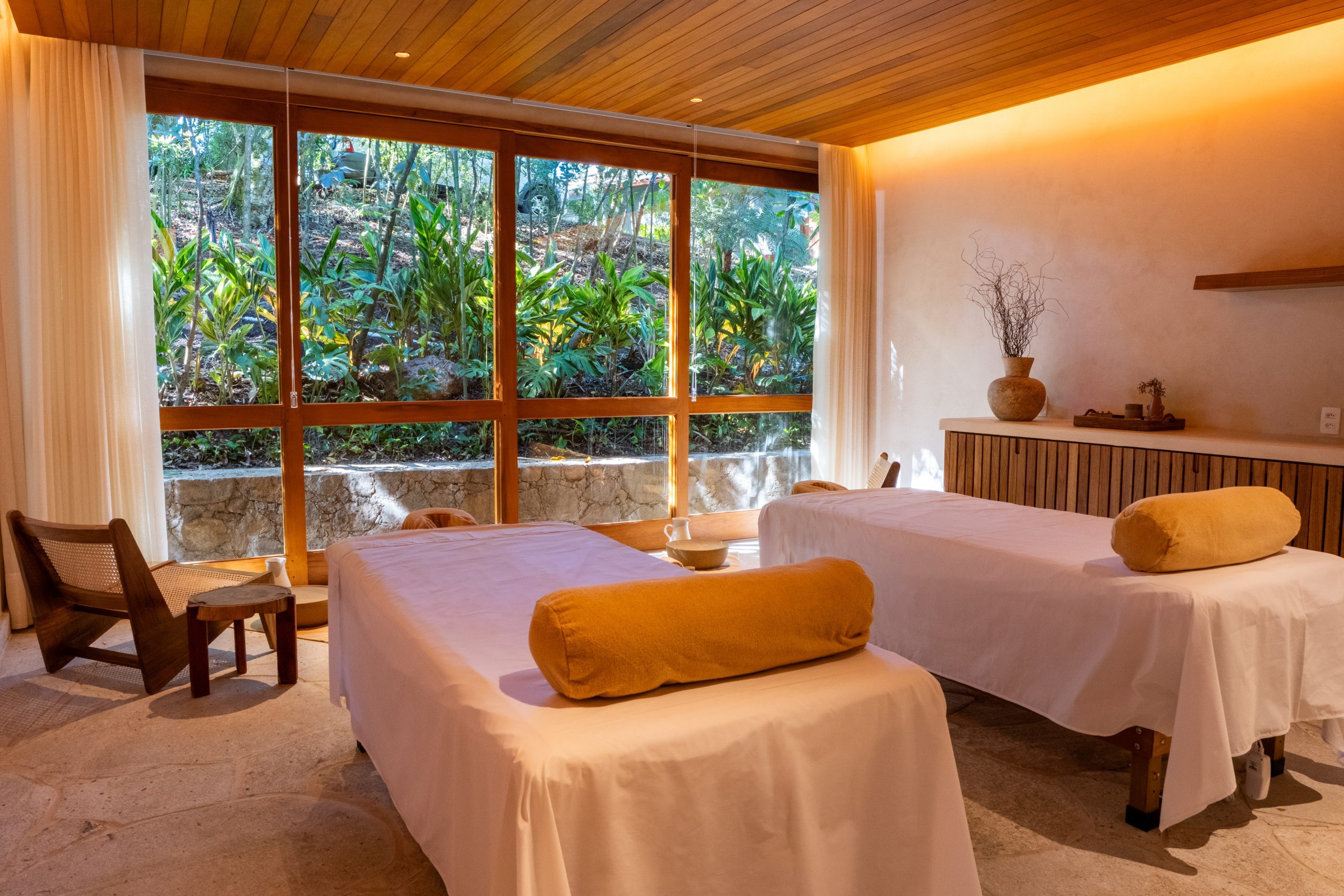 Two massage tables with rolled towels in a tranquil spa room, warm lighting, and large windows facing greenery.