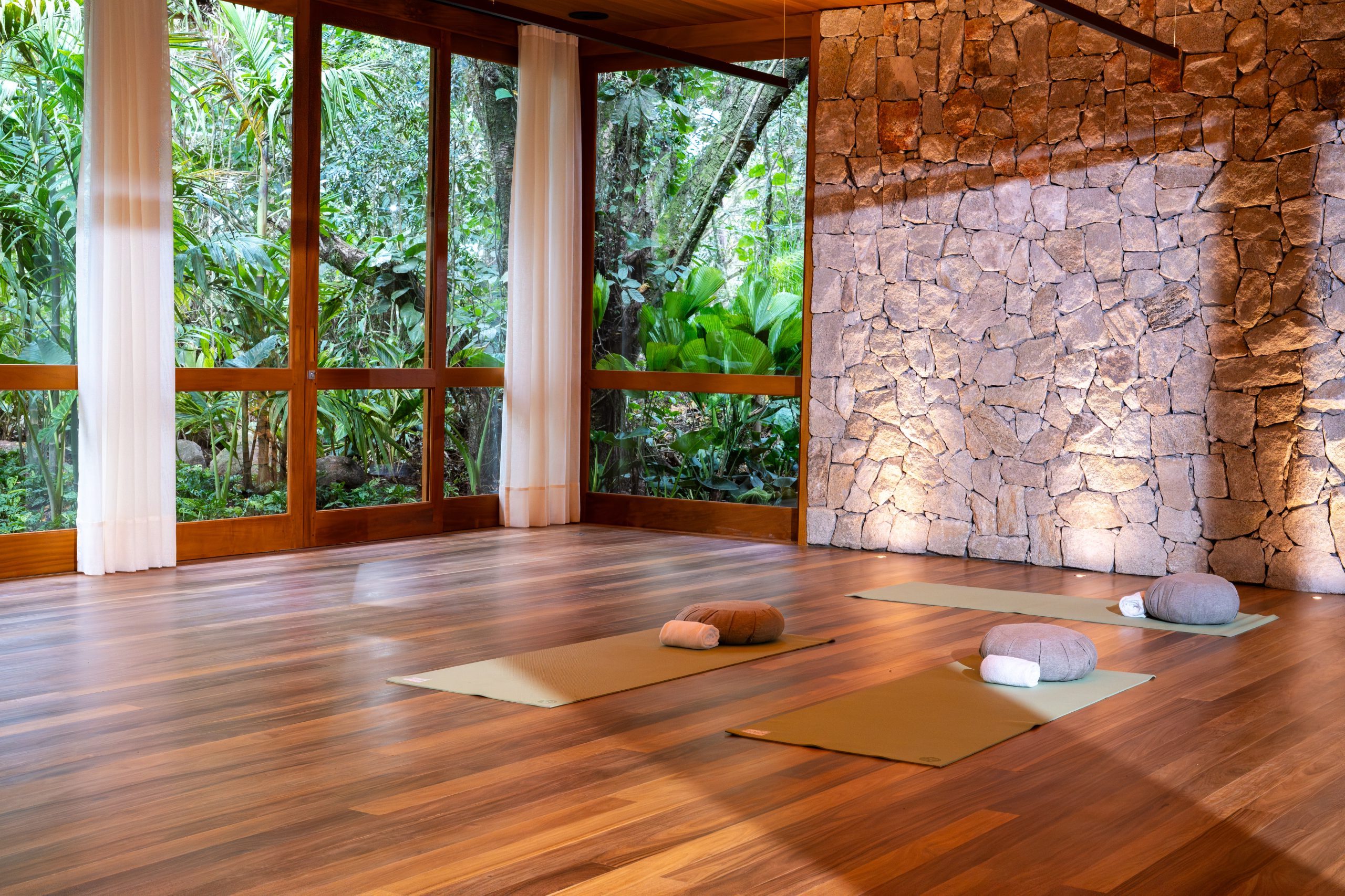 Serene yoga studio with wooden floors, stone wall, yoga mats, and lush green forest views through large windows.