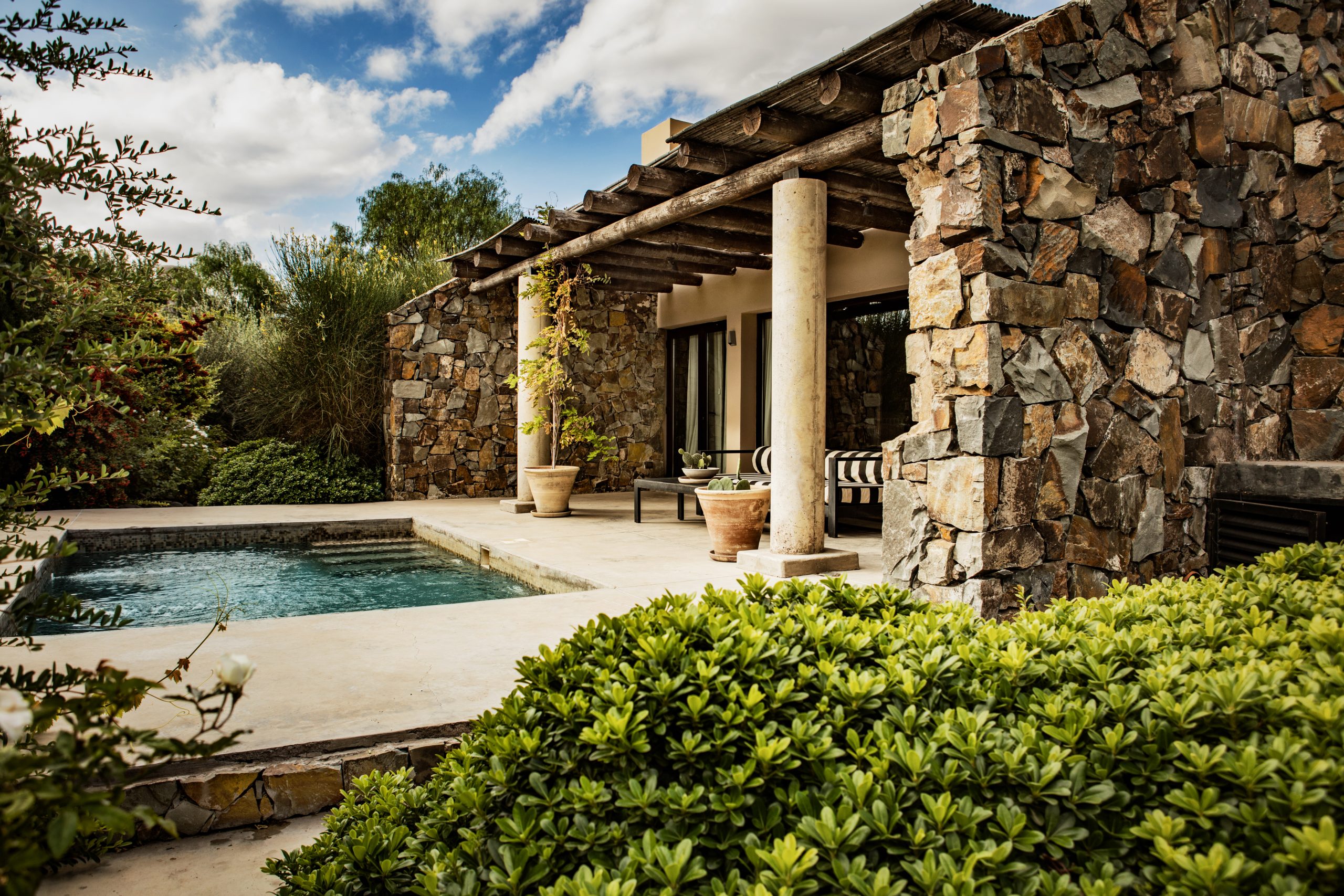 Stone villa with covered patio, pool, and lush garden, featuring rustic beams, columns, and outdoor seating.