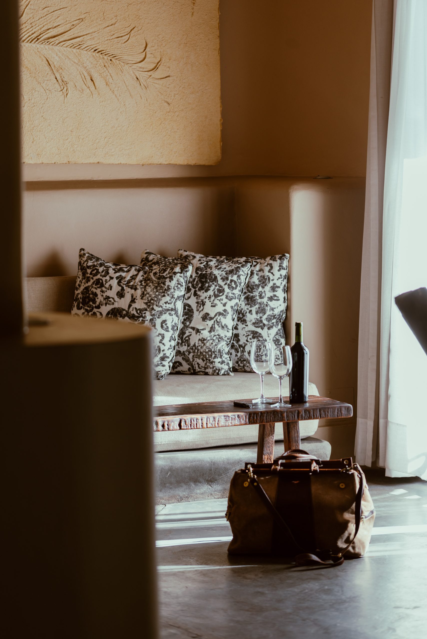 Cozy living area with patterned pillows, wooden table, wine bottle and glasses, and a leather bag near a sunlit window.