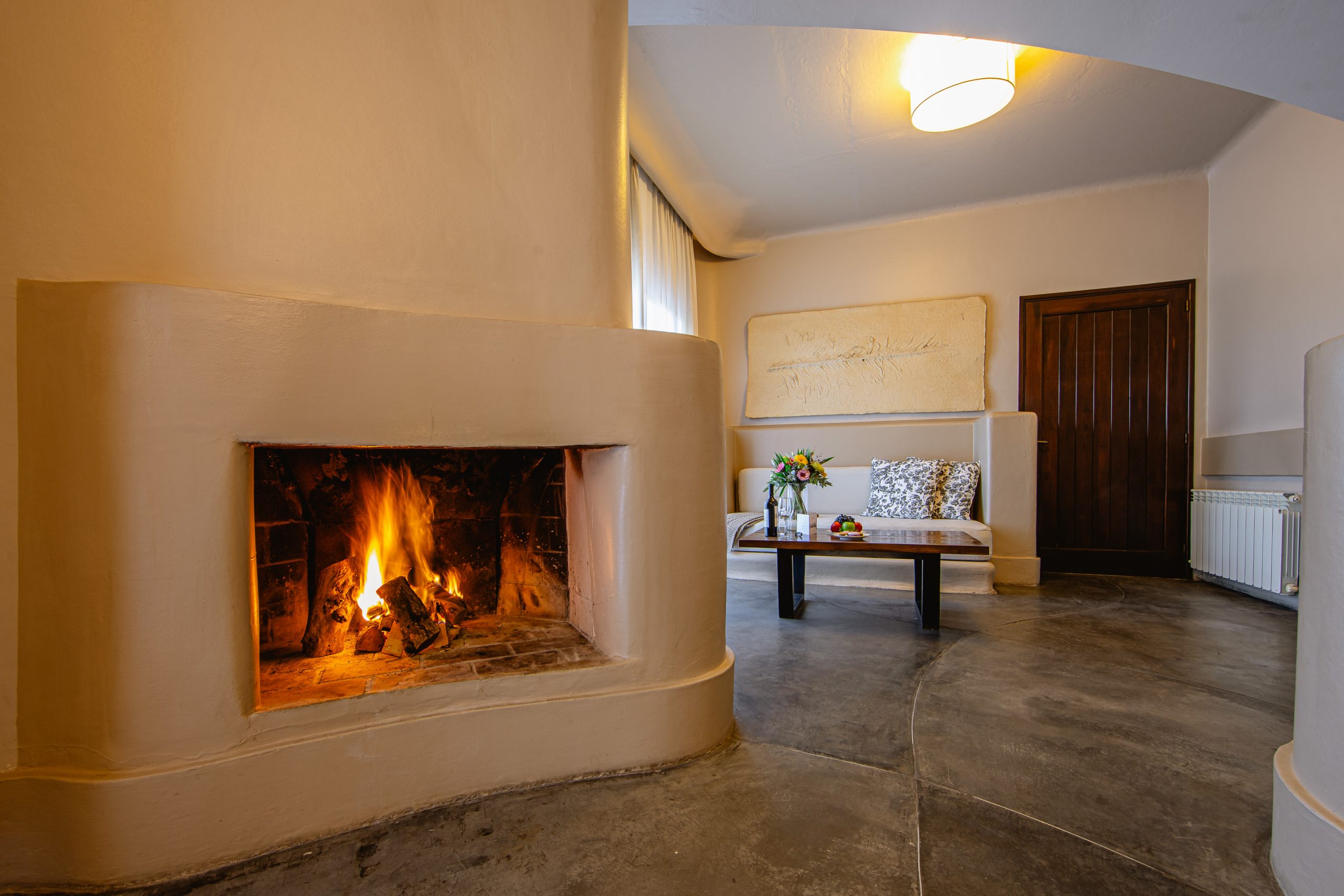 Cozy minimalist living room with curved plaster fireplace, lit fire, bench seating, wooden table, and warm neutral tones.