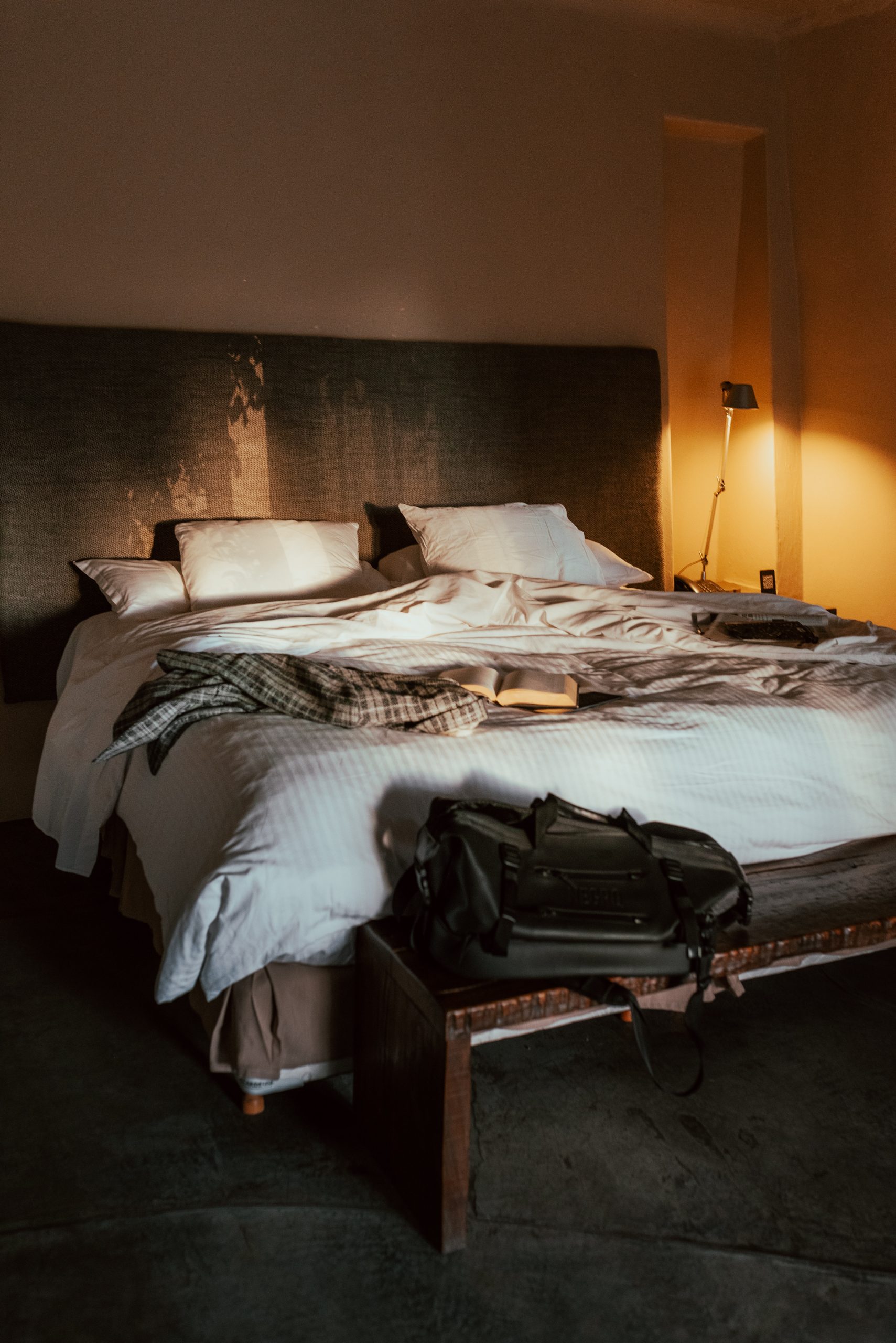 Warm-lit hotel bedroom with unmade bed, open book, bedside lamp, and travel bag on a bench.