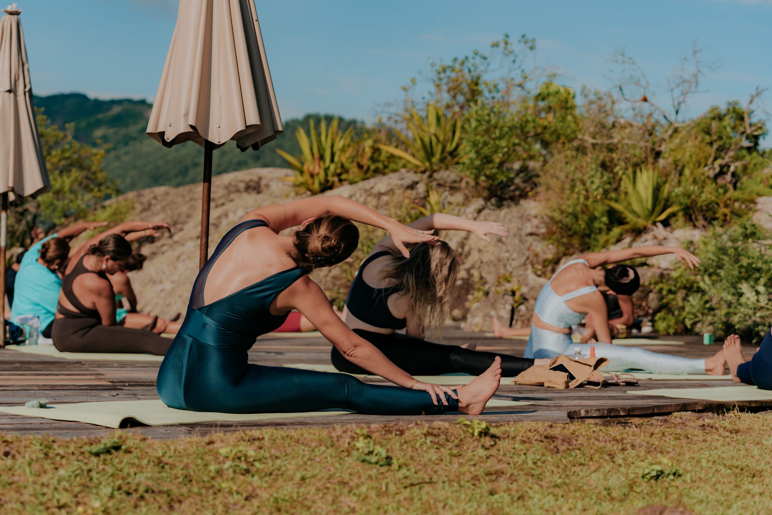 Group of women practicing outdoor yoga stretches on mats in a scenic hillside setting.