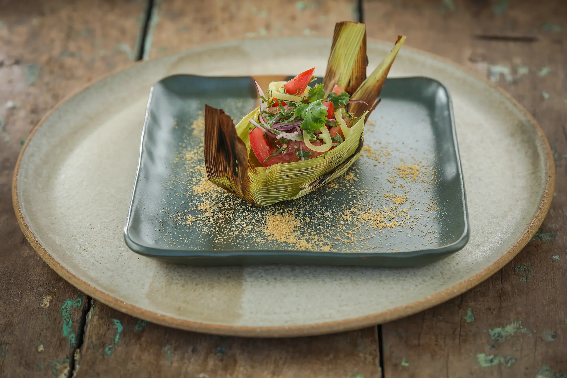 Artfully plated vegetable dish wrapped in charred leaves with herbs and seasoning on a square plate.
