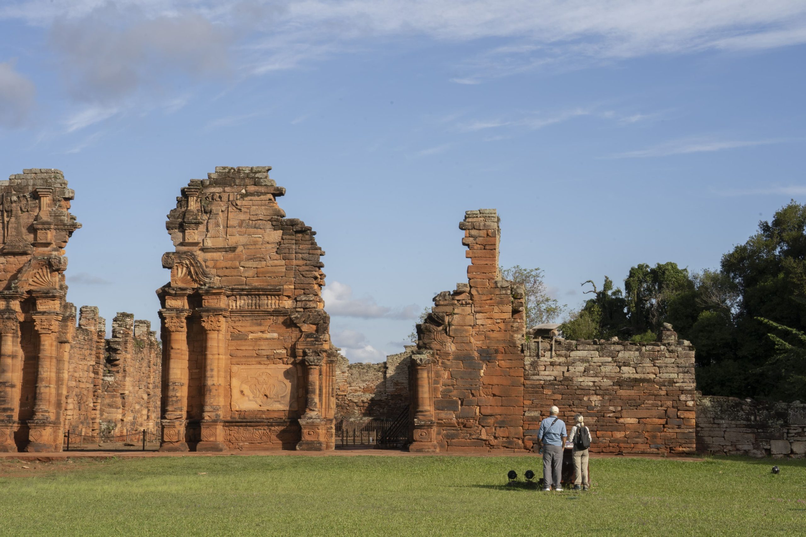 Two visitors stand on grass facing ancient red stone ruins with carved columns under a blue sky.