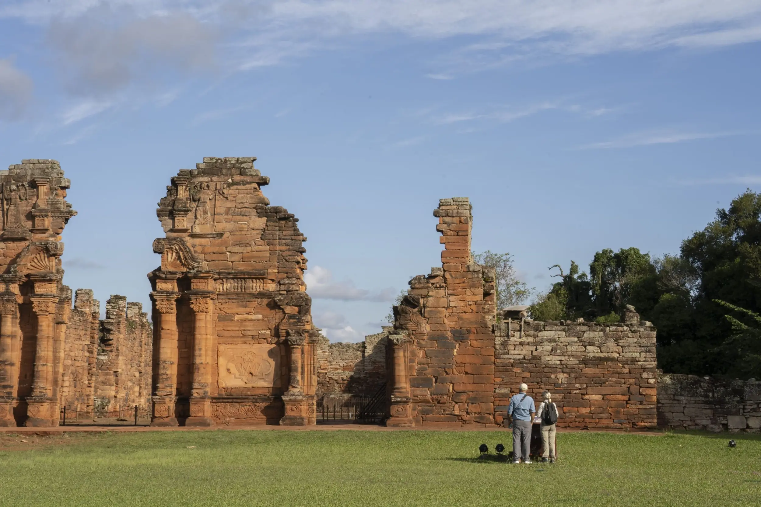 Two Visitors Stand On Grass Facing Ancient Red Stone Ruins With Carved Columns Under A Blue Sky.