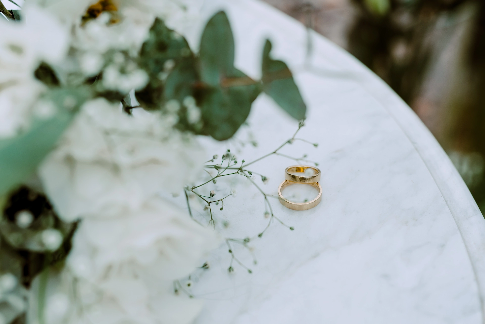 Two gold wedding rings on white marble beside a delicate bouquet of white flowers and greenery.