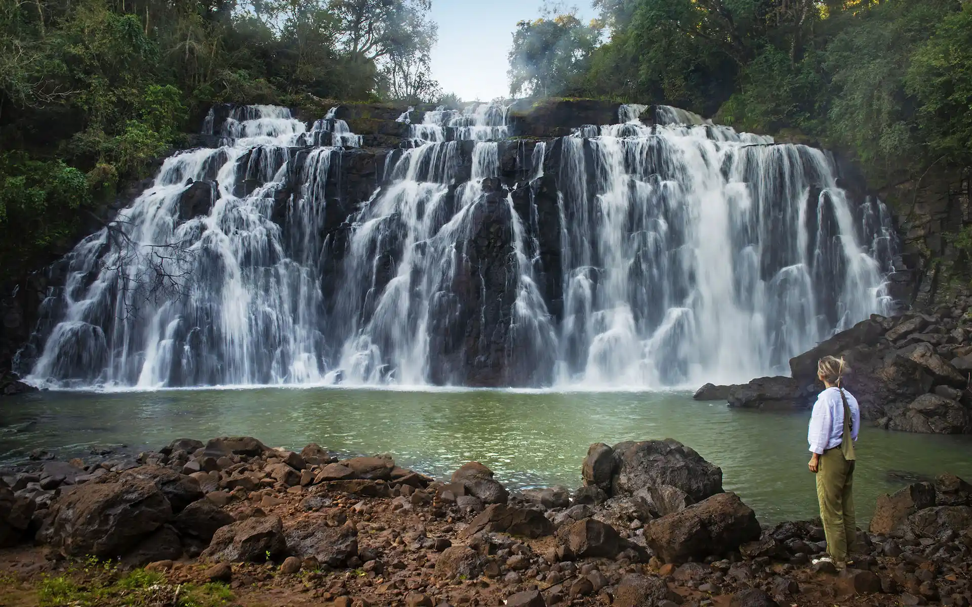 Wide waterfall cascading into a pool, with a person standing on rocky ground, surrounded by dense green forest.