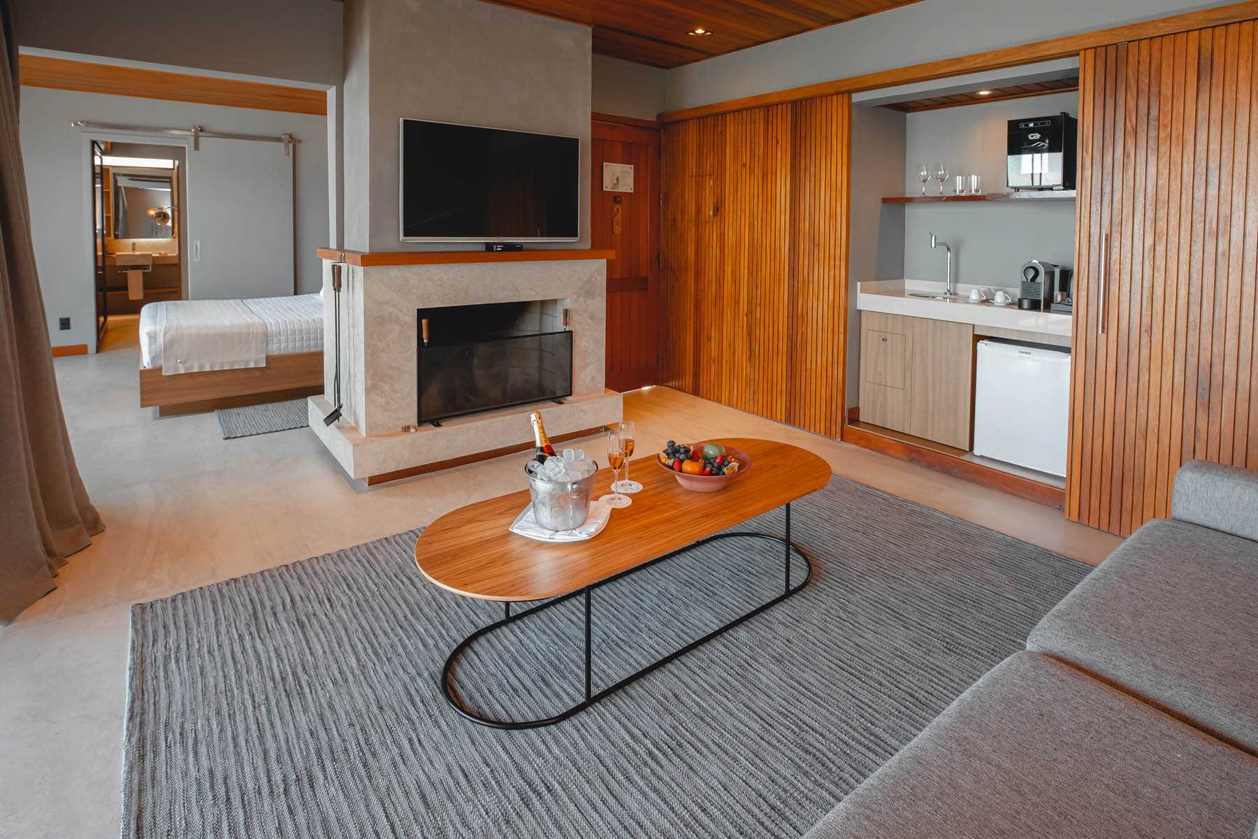 Modern hotel suite with fireplace, wall-mounted TV, kitchenette, coffee table with champagne, and bedroom visible in the background.
