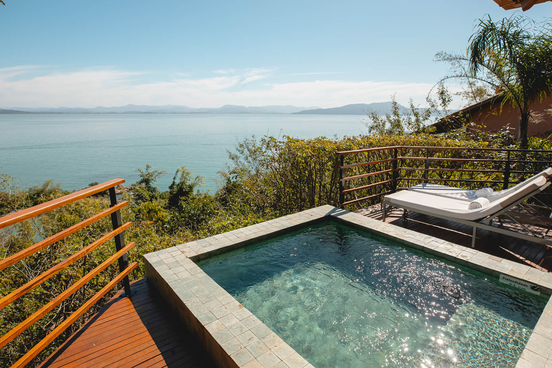 Private plunge pool on a wooden deck overlooking calm blue water, greenery, and distant hills under a clear sky.