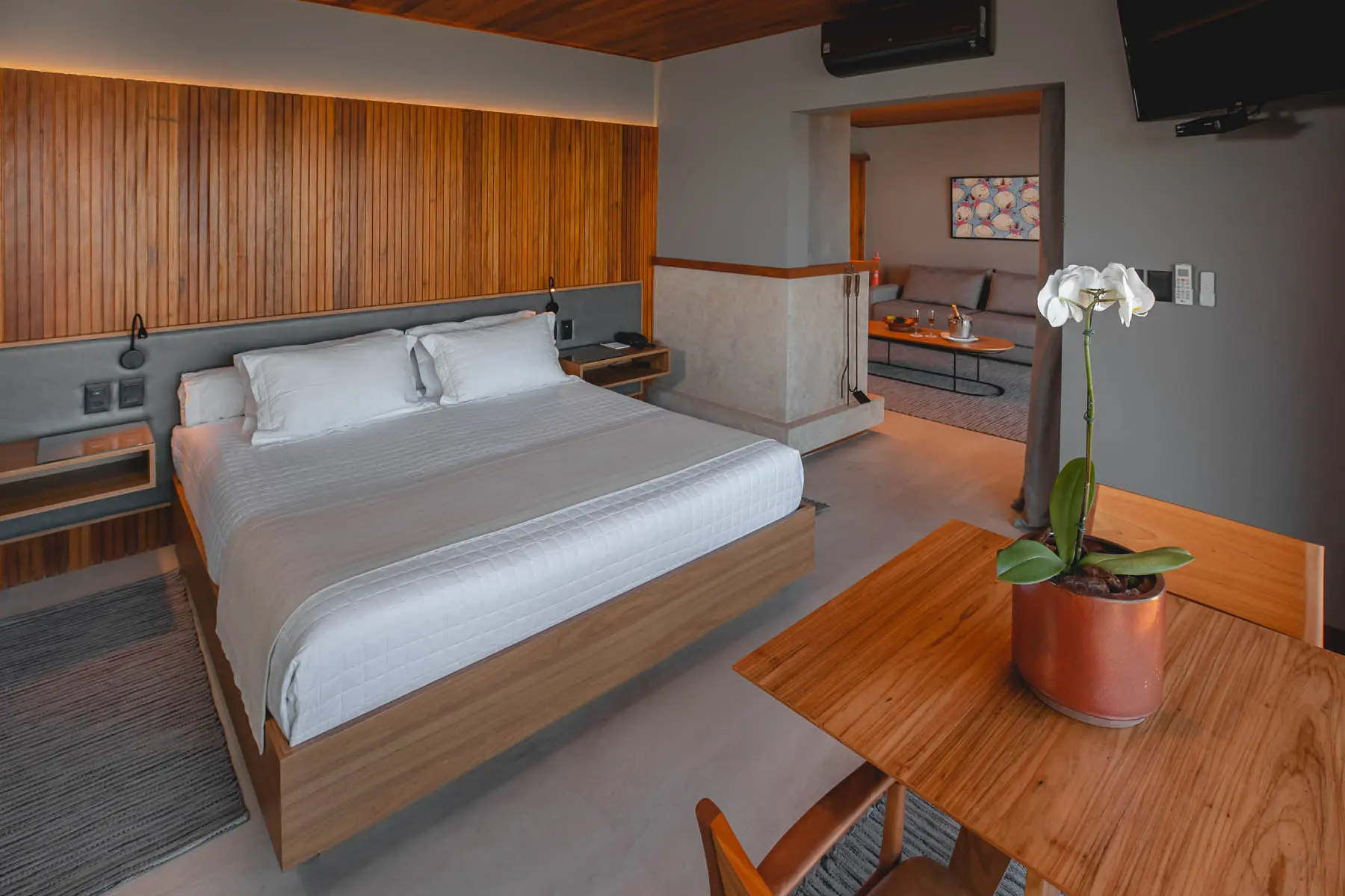 Modern Hotel Bedroom With Wooden Paneling, Queen Bed, Minimalist Furnishings, And An Open Doorway To A Cozy Lounge Area.