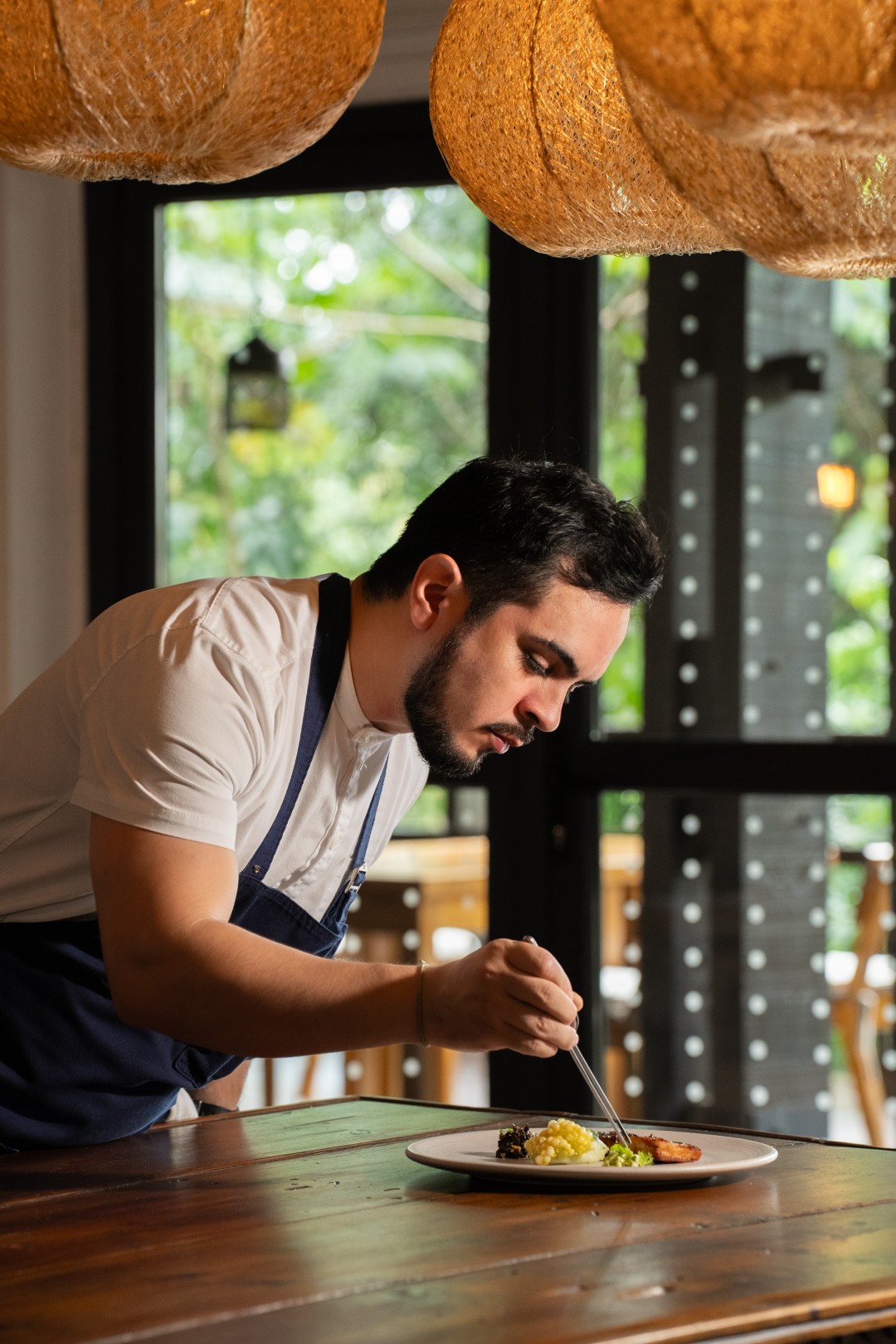 Chef carefully plating a gourmet dish on a table, focusing on detail inside a softly lit restaurant with natural decor.