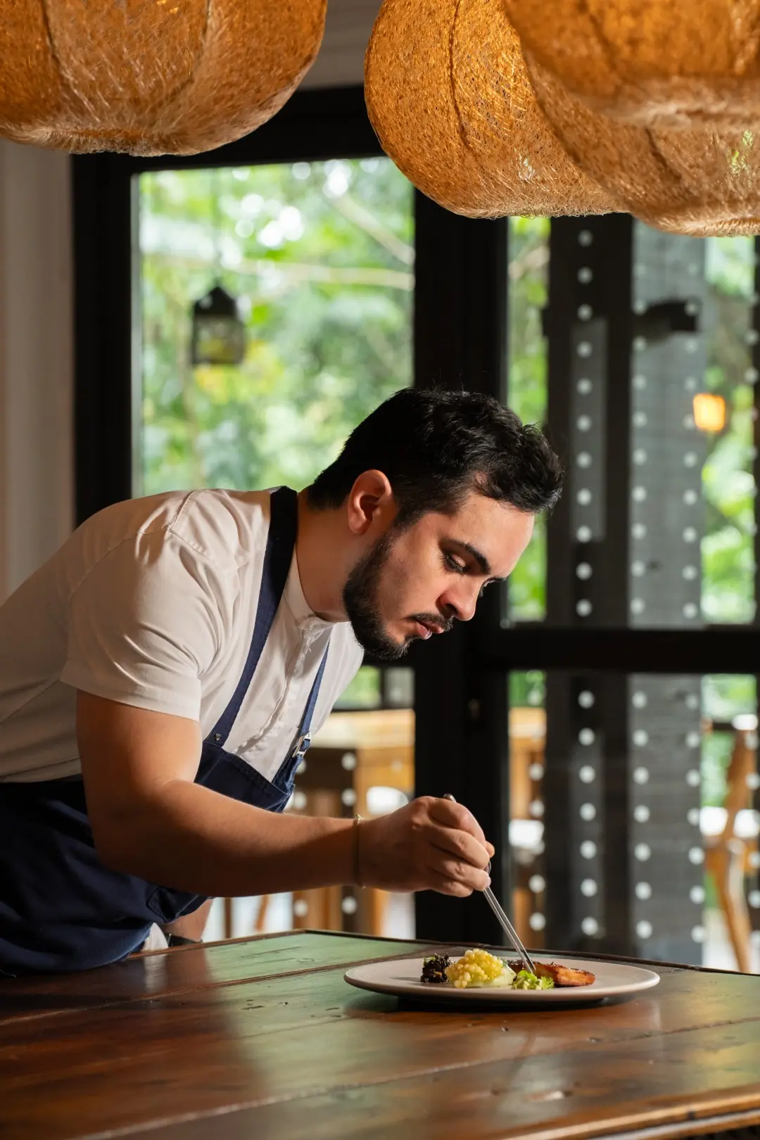 Chef Carefully Plating A Gourmet Dish On A Table, Focusing On Detail Inside A Softly Lit Restaurant With Natural Decor.