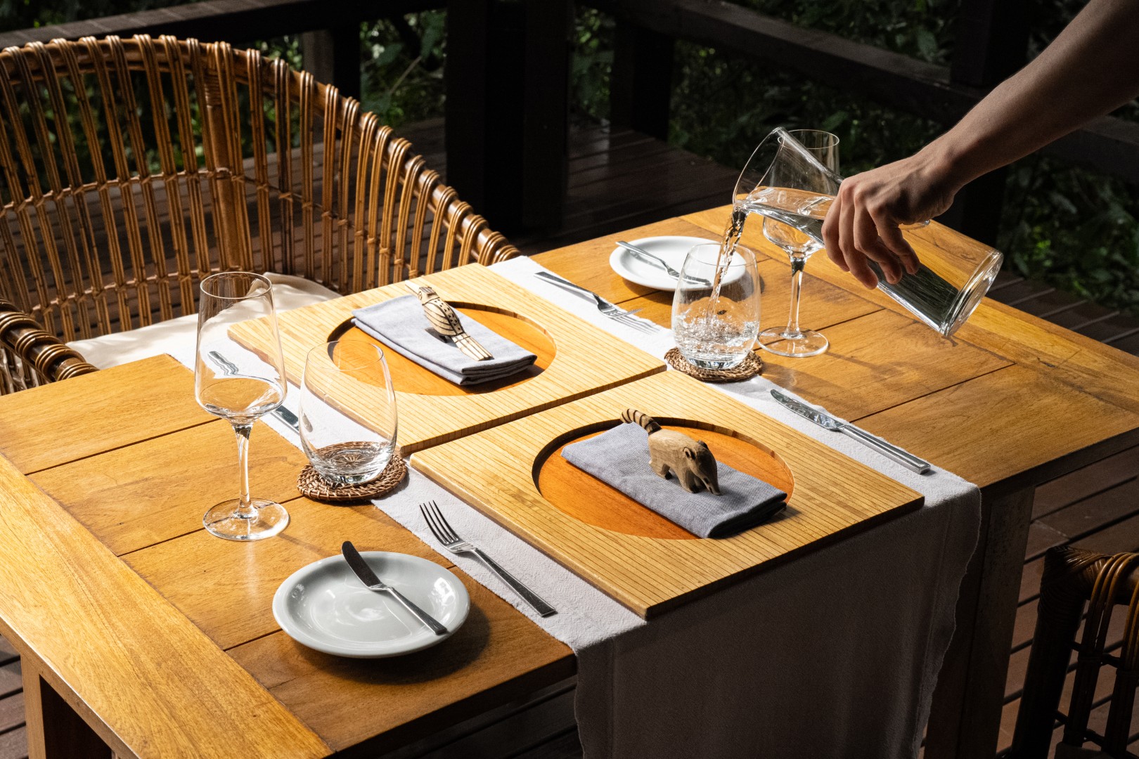 Outdoor dining table set with wooden plates, wine glasses, and napkins as water is poured into a glass.