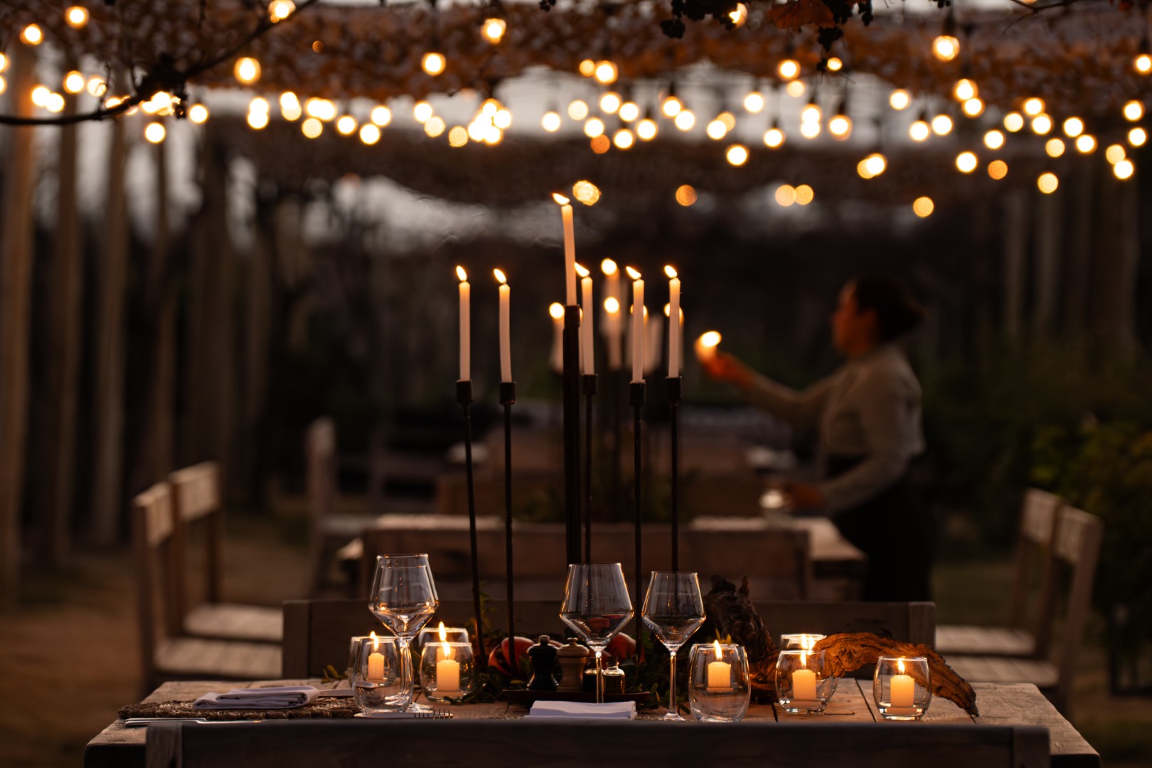 Candlelit outdoor dining table with wine glasses and string lights overhead, creating a warm, intimate evening atmosphere.
