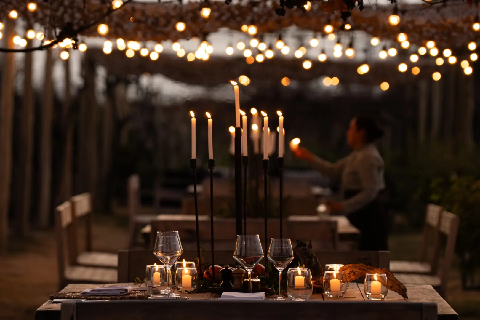 Candlelit Outdoor Dining Table With Wine Glasses And String Lights Overhead, Creating A Warm, Intimate Evening Atmosphere.