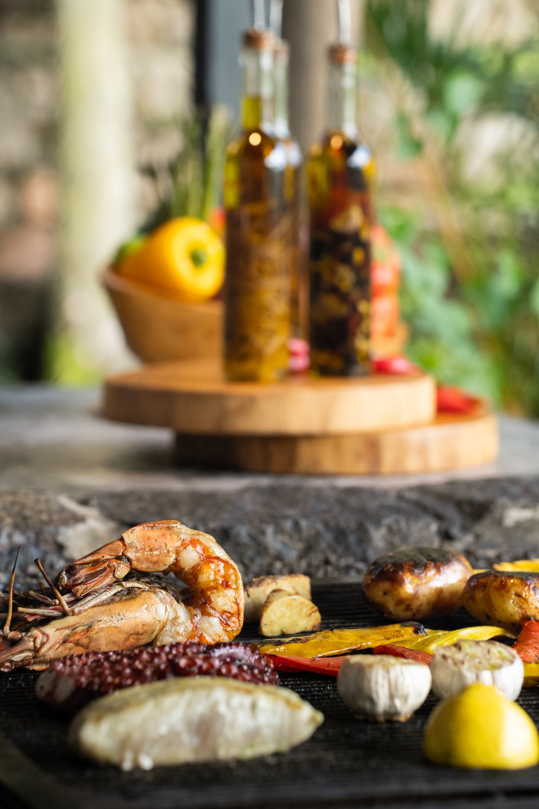Grilled seafood platter with shrimp, fish, potatoes, vegetables, and lemon, with olive oil bottles blurred in background.
