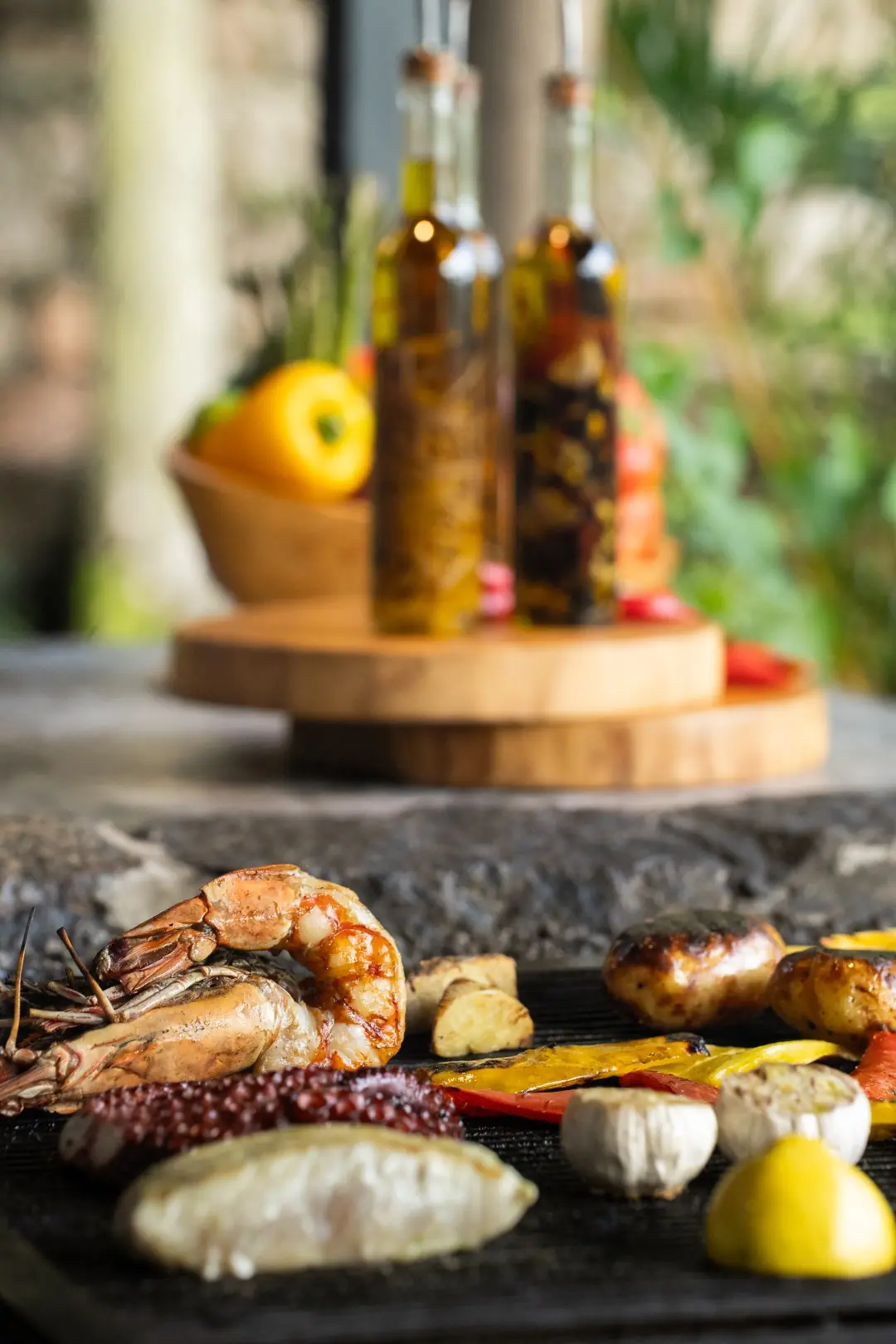 Grilled Seafood Platter With Shrimp, Fish, Potatoes, Vegetables, And Lemon, With Olive Oil Bottles Blurred In Background.