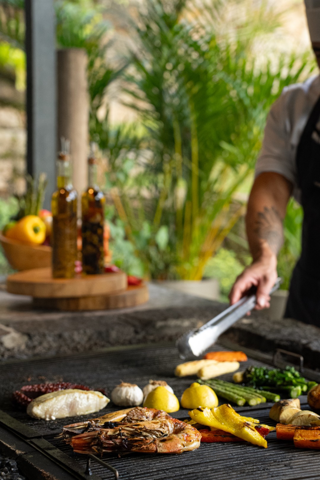 Chef grilling seafood and vegetables outdoors, with shrimp, fish, lemons, asparagus, and herbs on a hot grill.