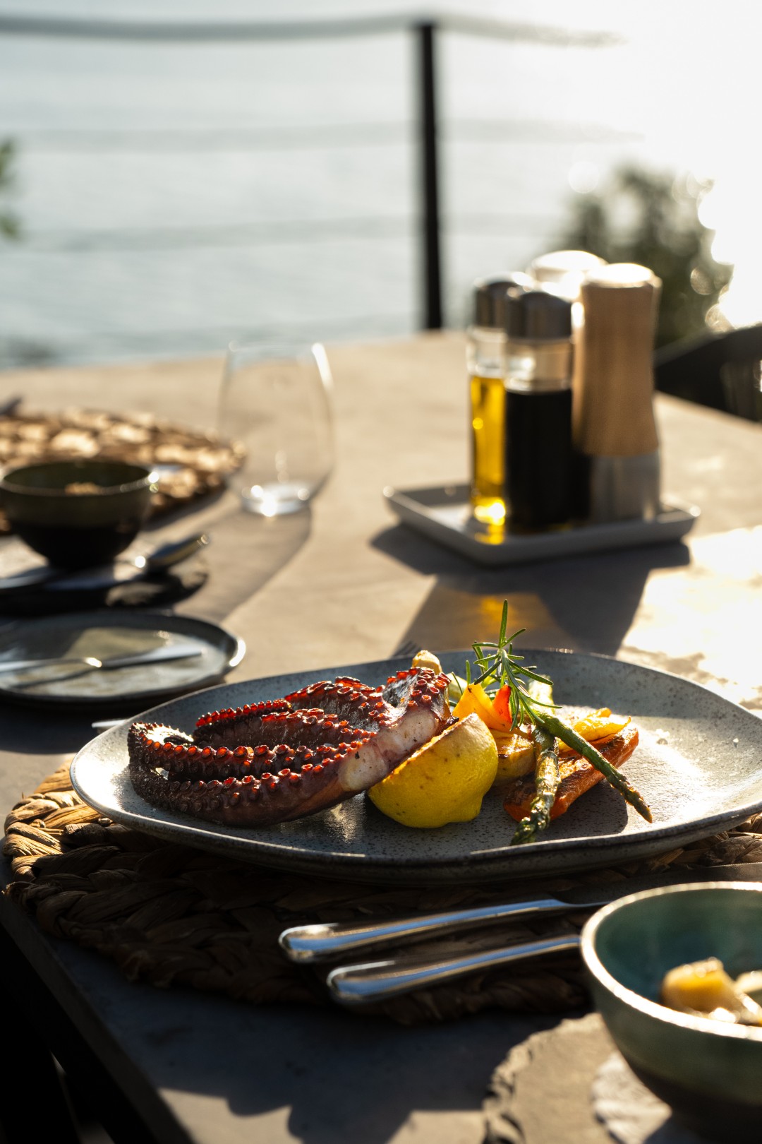 Grilled octopus with lemon, vegetables, and herbs served on a plate at an outdoor seaside table in warm sunlight.