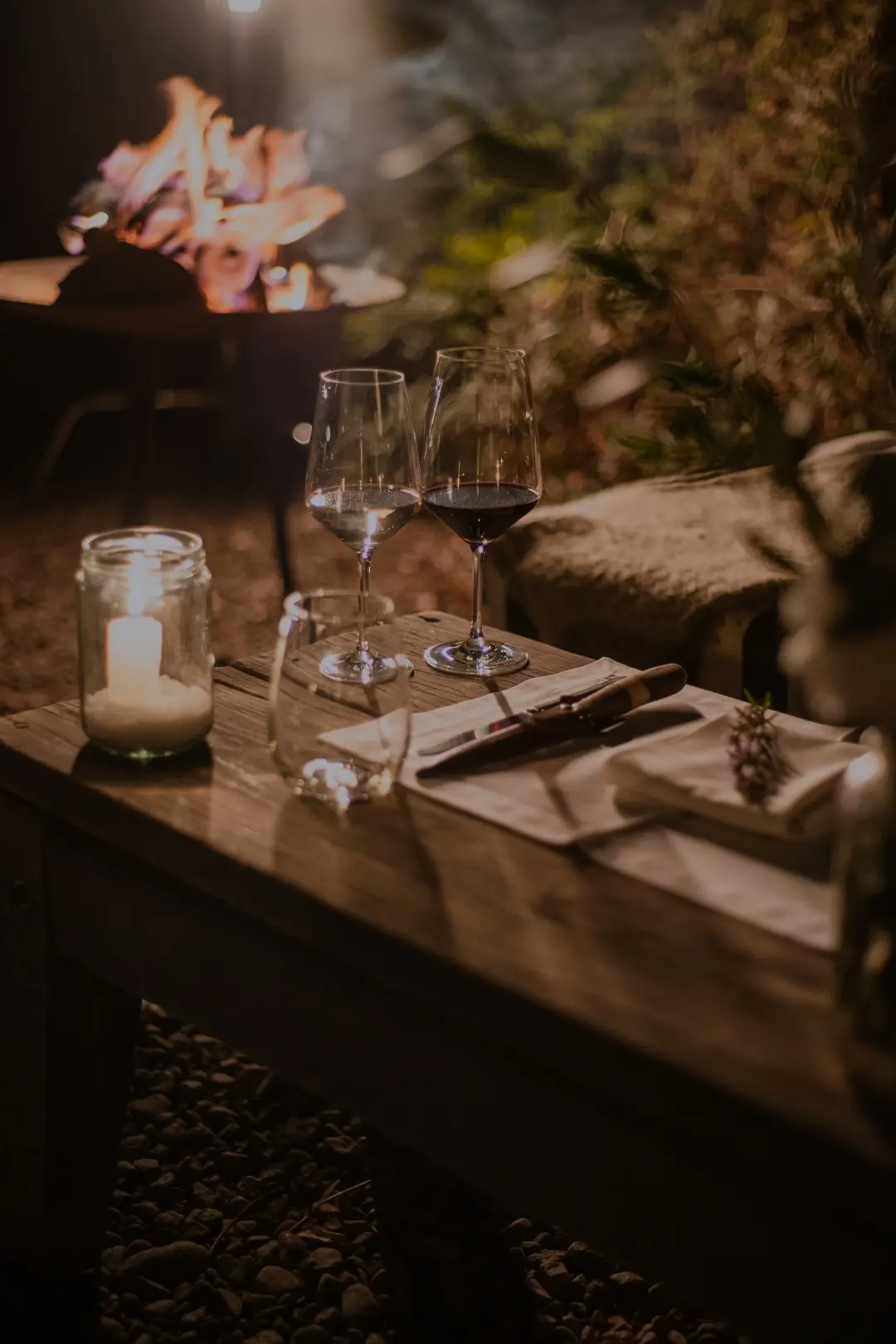 Candlelit outdoor table with wine glasses and place setting beside a glowing fire pit at night.