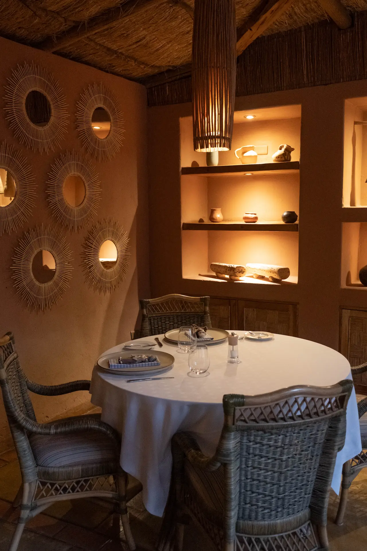 Intimate dining nook with round table, woven chairs, warm lighting, textured walls, and decorative shelves with pottery.