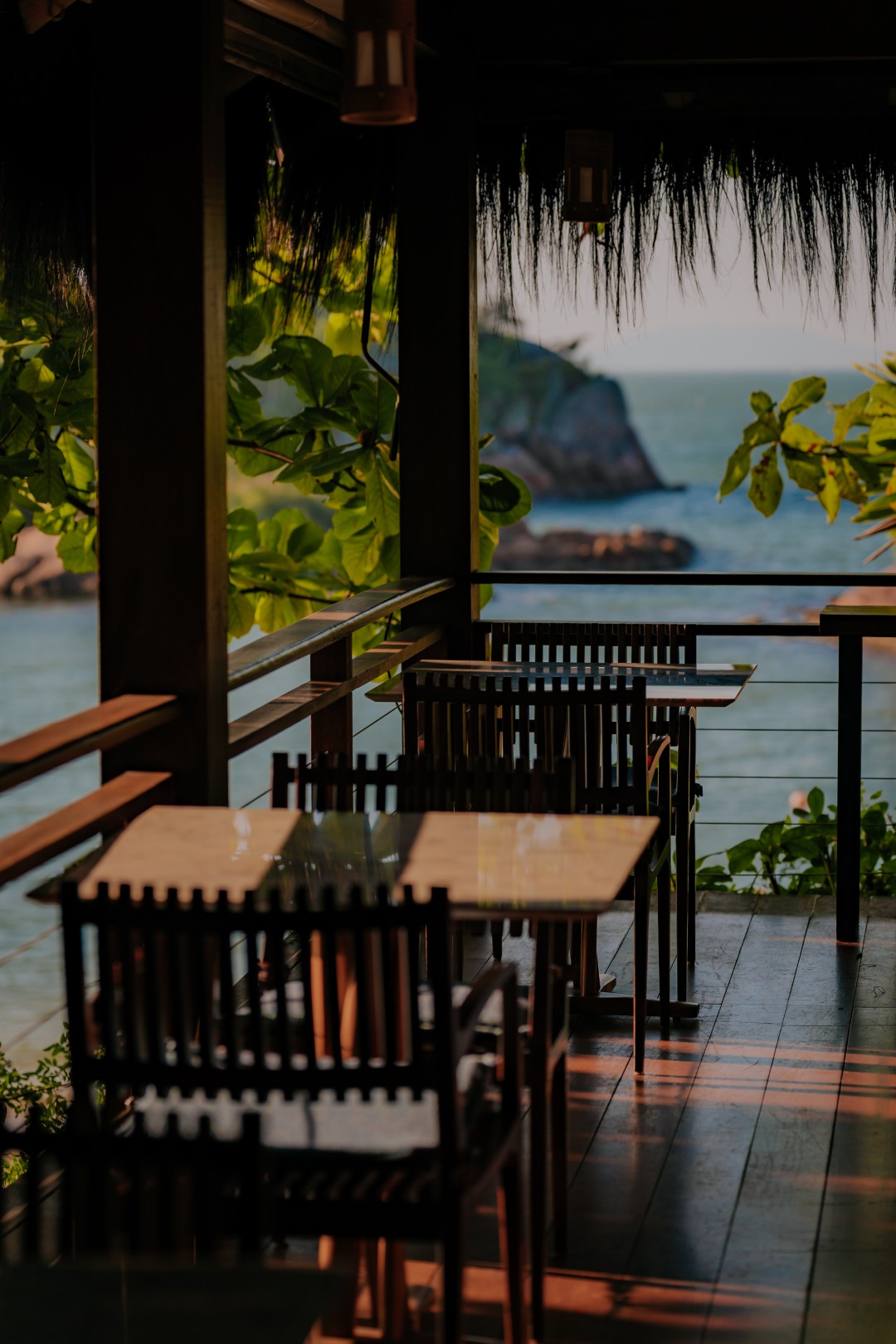 Outdoor restaurant terrace with wooden tables and chairs overlooking the ocean, framed by tropical greenery.