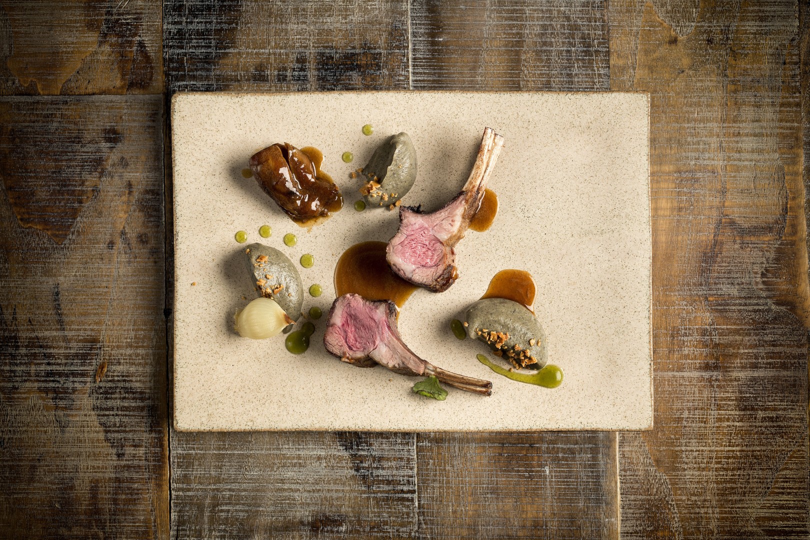 Artfully plated lamb chops with purée, sauce, and garnishes on a rectangular ceramic plate set on a rustic table.