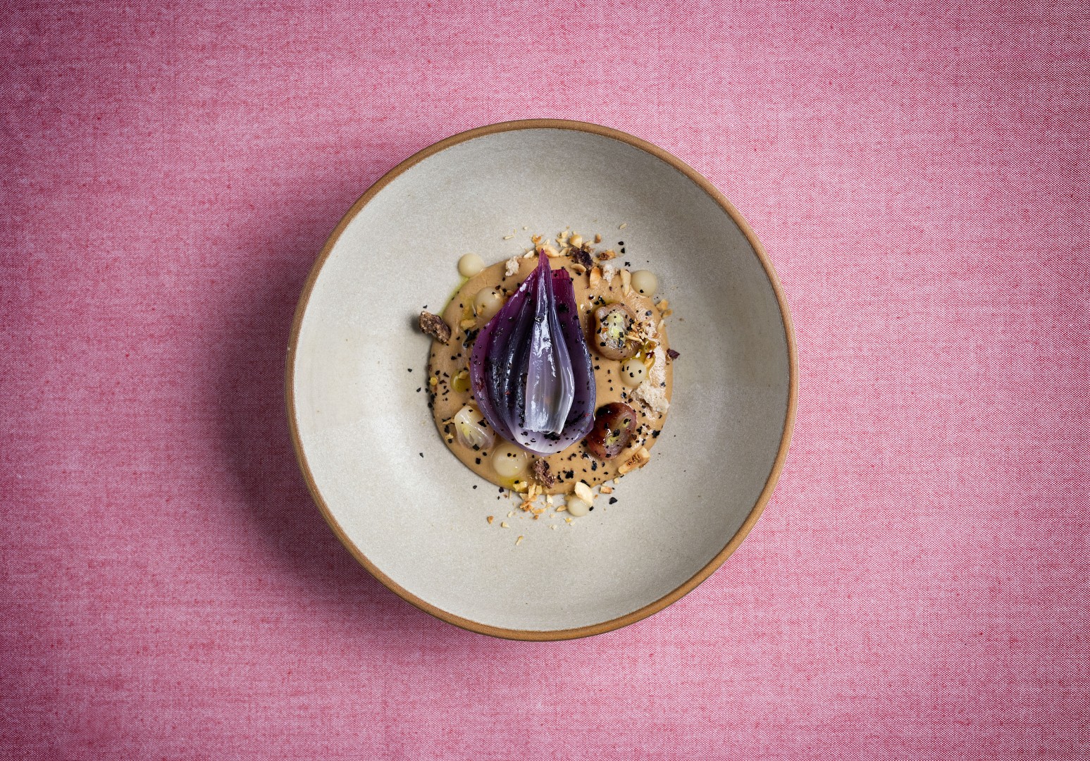 Artfully plated gourmet dish with roasted onion, sauce, and garnishes centered in a ceramic bowl on a pink backdrop.