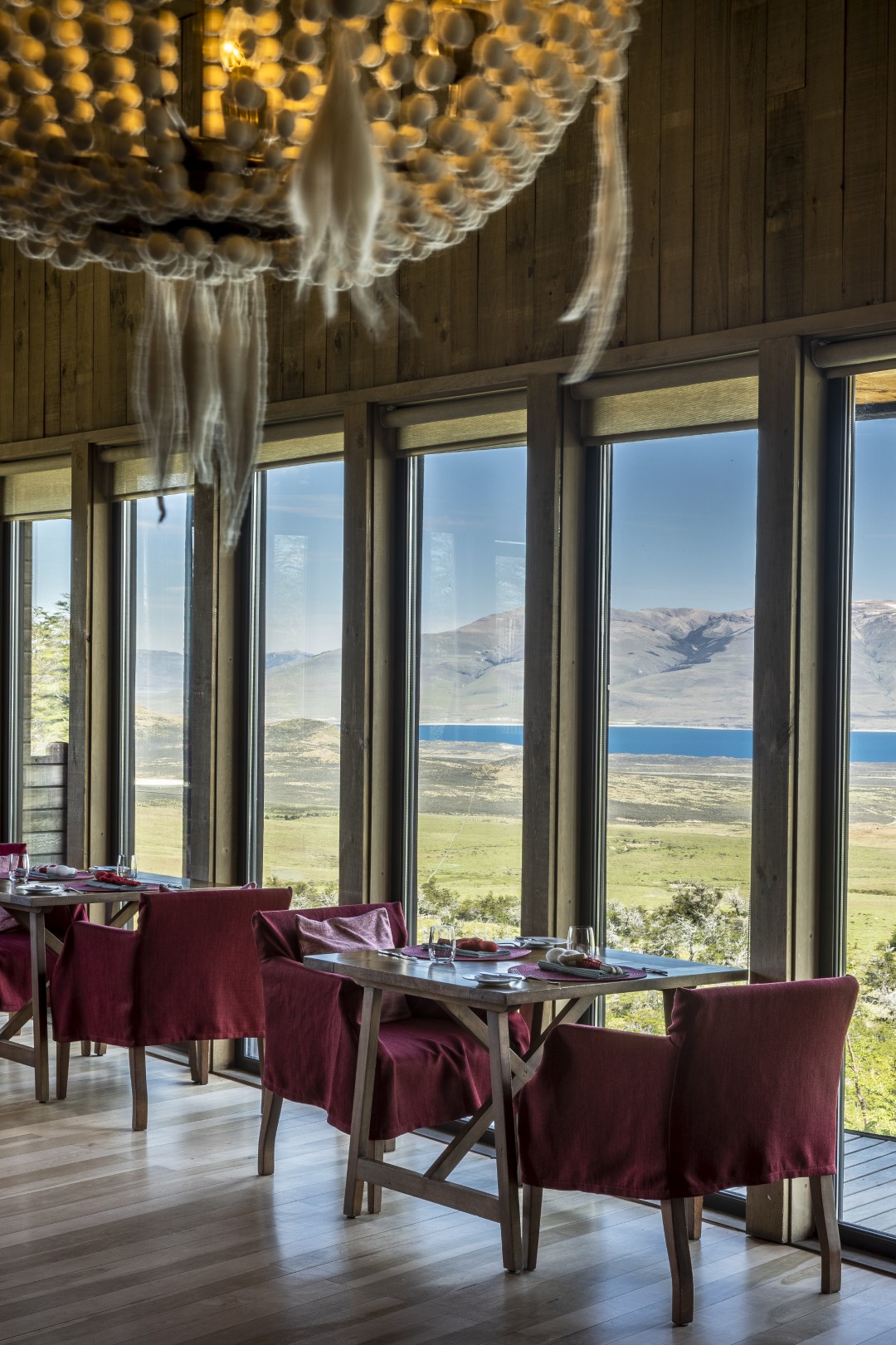 Elegant dining area with red chairs, wooden tables, large windows, and sweeping views of grasslands, lake, and mountains.