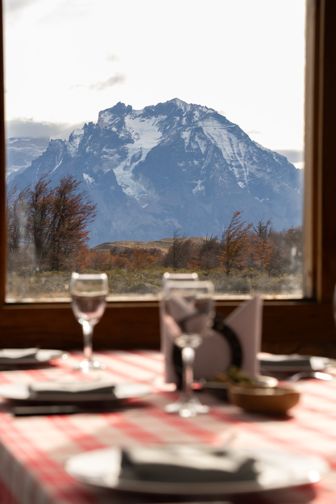 Dining table set by a window with wine glasses, overlooking a snow-capped mountain and autumn landscape.