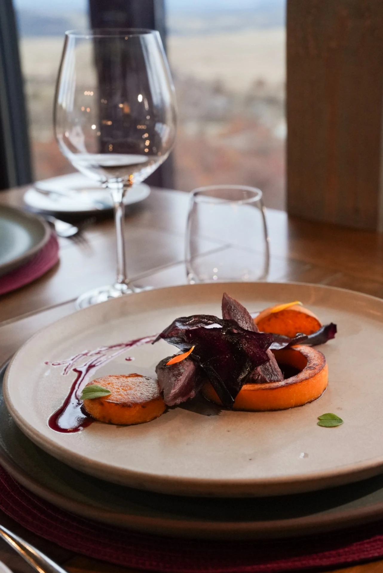 Plated gourmet dish with roasted vegetables and sauce on a ceramic plate, set on a dining table by a window.