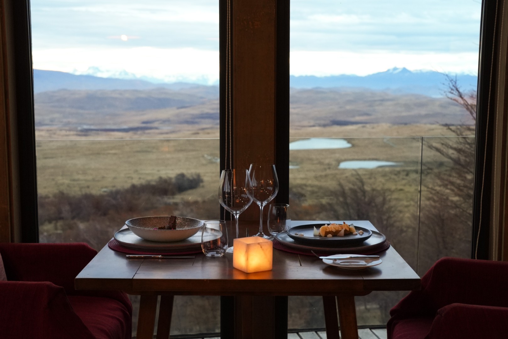 Candlelit dining table for two by a window, overlooking open plains and distant mountains at dusk.