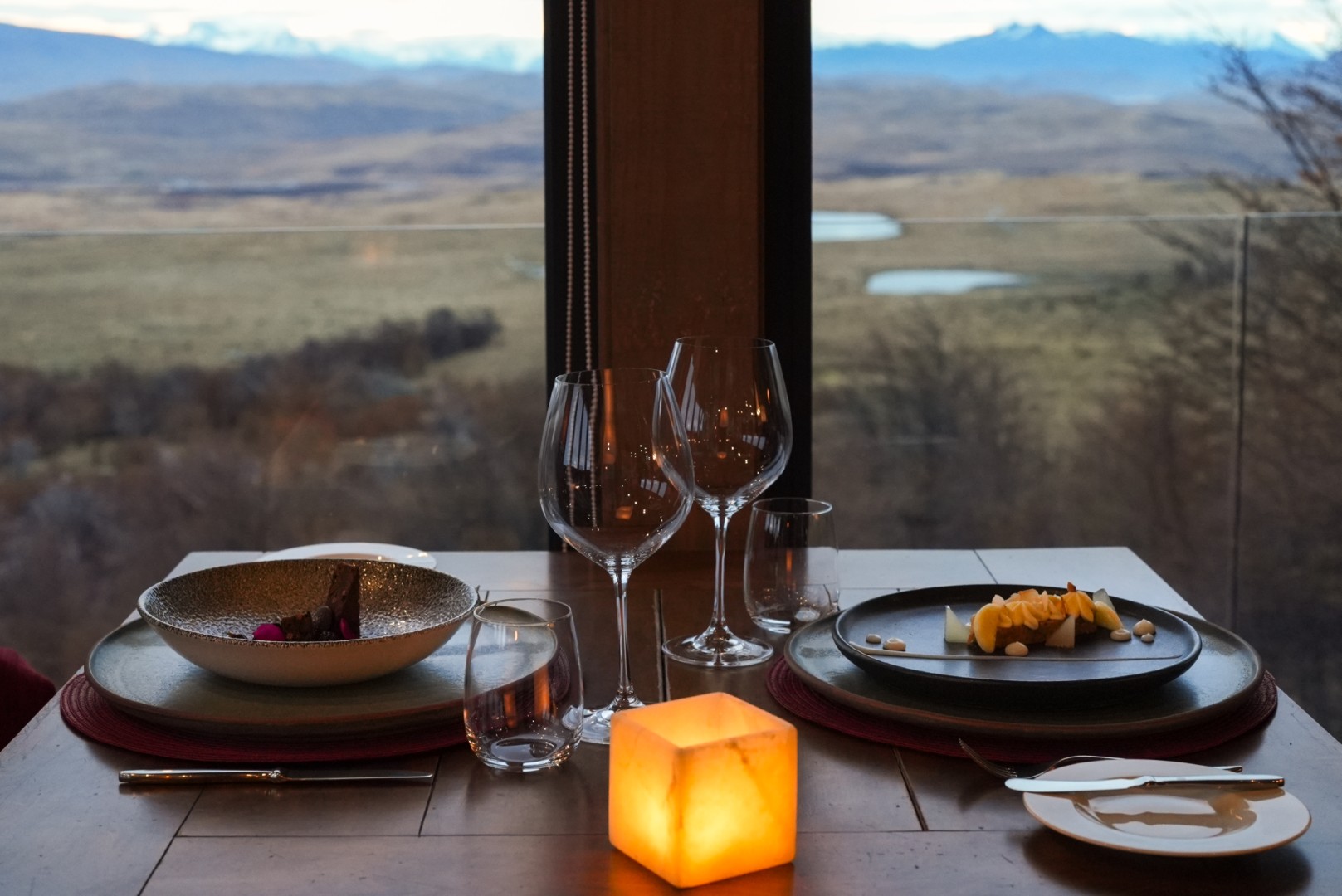 Candlelit dining table with plated dishes and wine glasses by a window overlooking open plains and distant mountains.