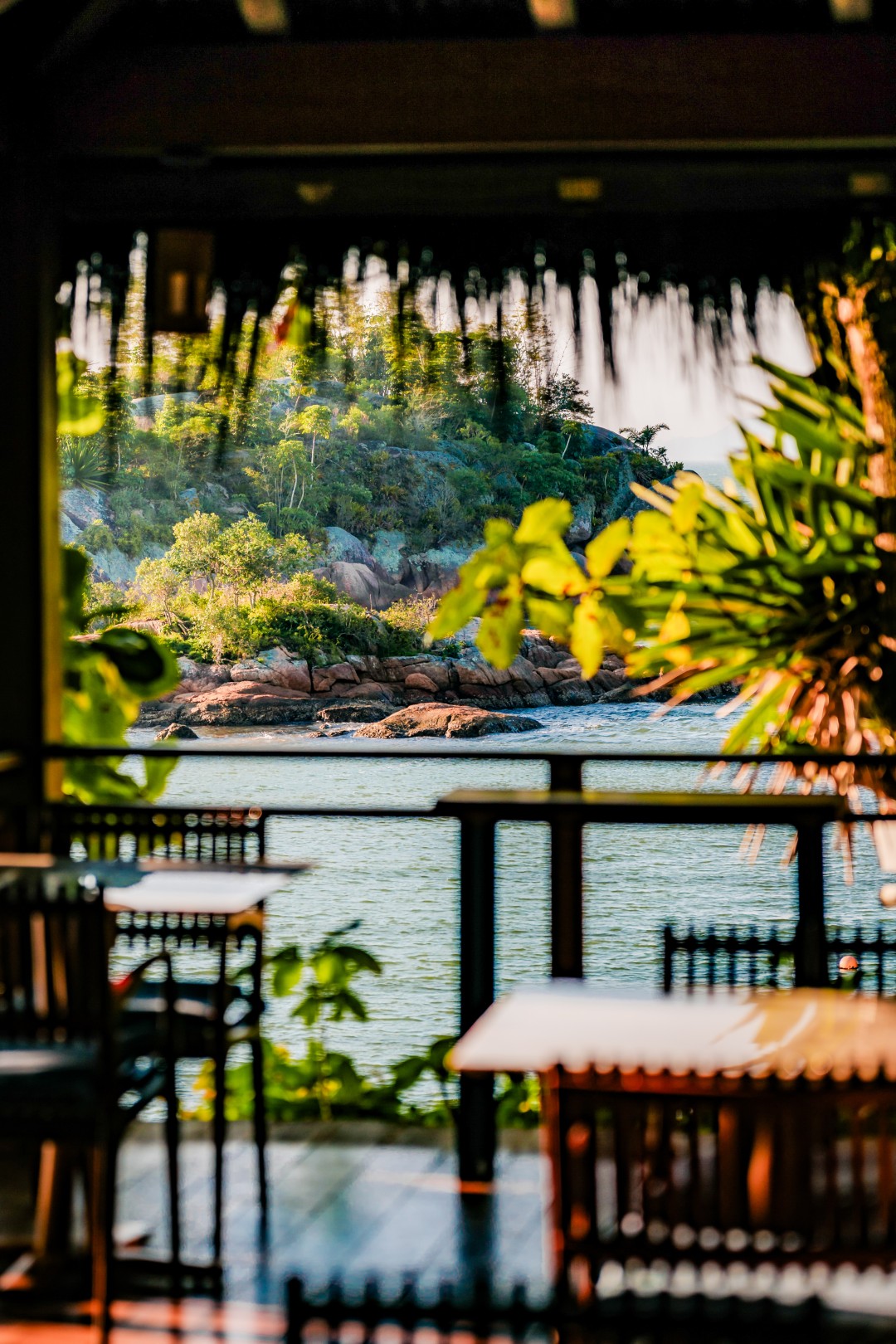 Open-air dining terrace framed by greenery, overlooking calm water and a rocky, tree-covered island.