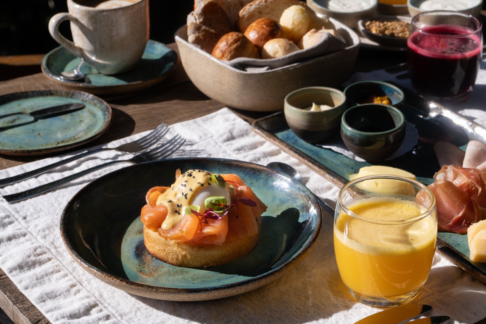 Breakfast spread with smoked salmon eggs Benedict, fresh bread, orange juice, coffee, and small bowls of condiments on a wooden table.