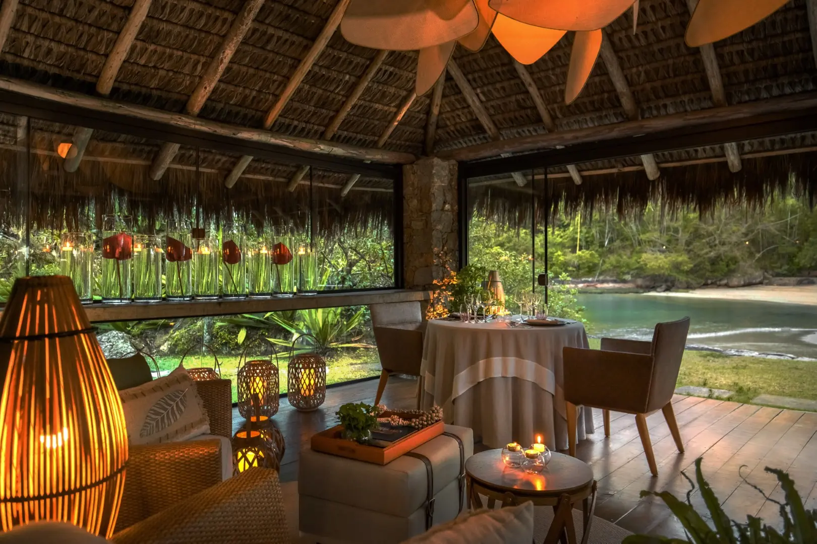 Romantic Open-Air Dining Room With Candlelit Table, Thatched Roof, And Lush Tropical Beach View.