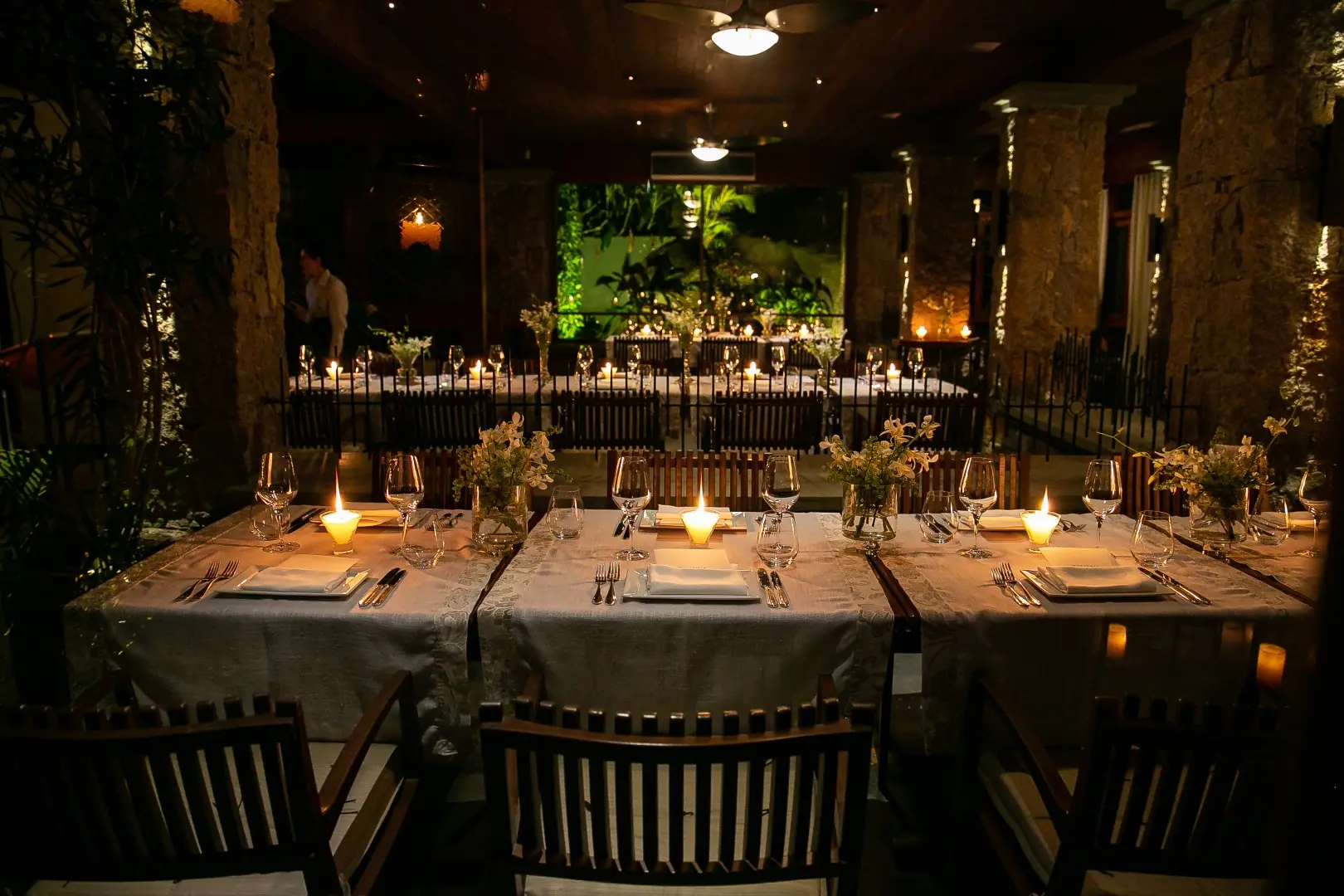 Elegant Candlelit Dining Room With Long Tables Set For Dinner, White Linens, Glassware, Flowers, And Warm Ambient Lighting.