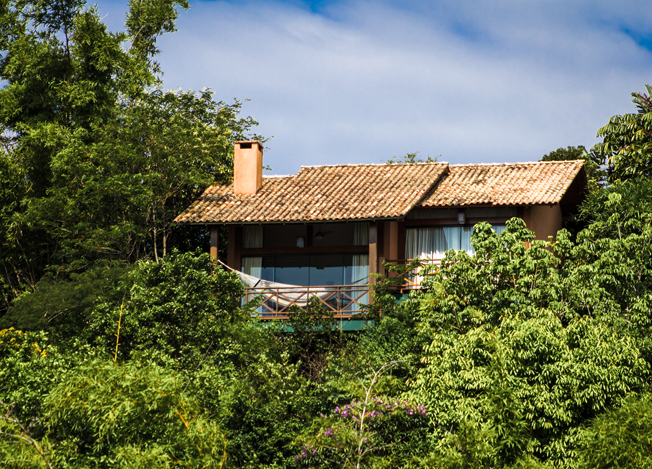 Secluded hillside cabin with a tiled roof and balcony, surrounded by dense green forest.