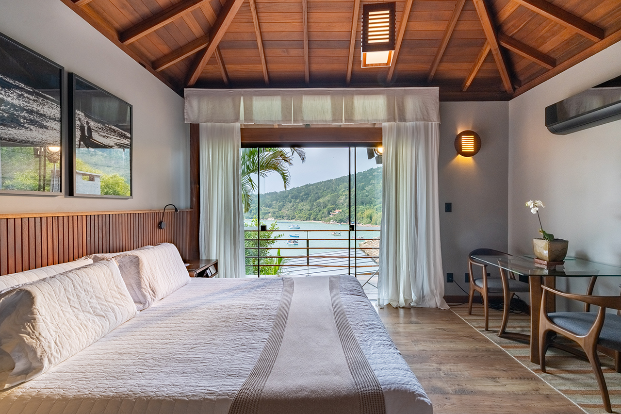 Bright bedroom with wooden ceiling, large bed, and glass doors opening to a balcony overlooking water and green hills.