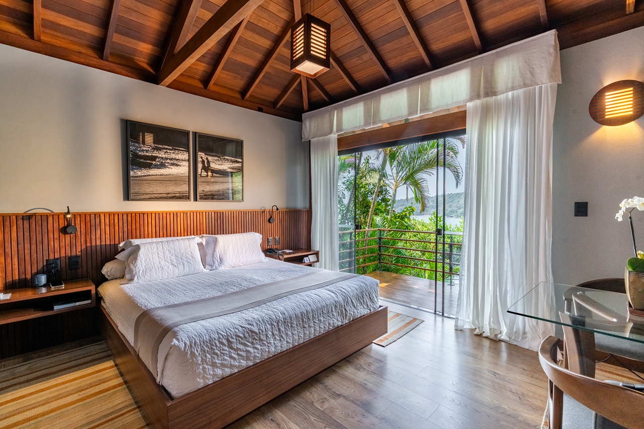 Modern bedroom with wooden ceiling, large bed, and sliding doors opening to a balcony with tropical views.